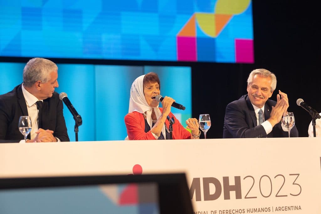 Una Madre de Plaza de Mayo, con el pañuelo blanco, habla al micrófono en la mesa de apertura mientras quienes la acompañan la escuchan y aplauden, ante la pantalla del FMDH2023.