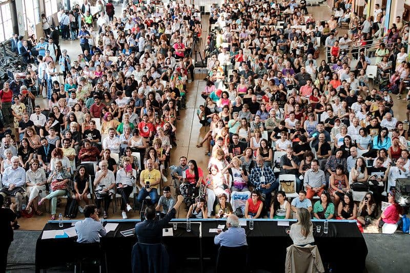 Vista desde arriba de un salón colmado: cientos de personas sentadas frente a la mesa de un panel.