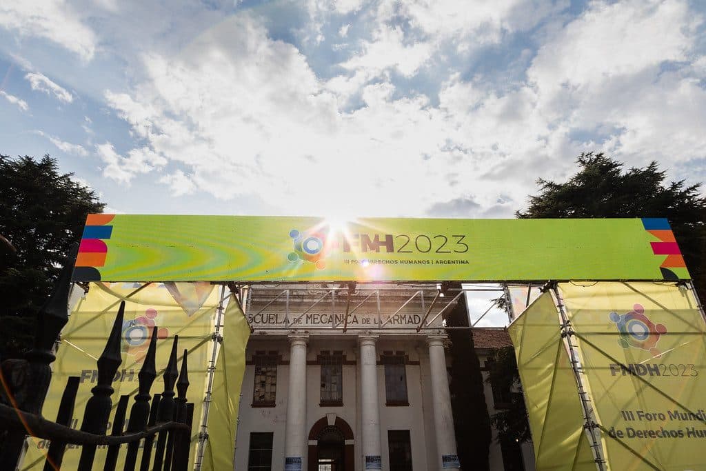 Pórtico con el banner FMDH2023 frente al edificio de la ex Escuela de Mecánica de la Armada, a contraluz bajo un cielo con nubes.