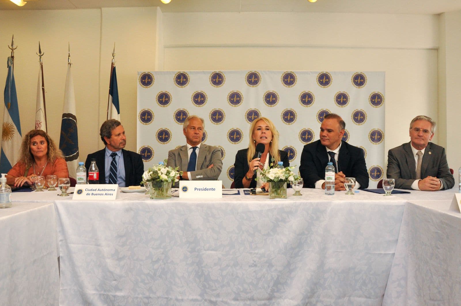 Mesa principal de un encuentro del notariado: una autoridad habla al micrófono junto a otras cinco, ante el telón del CFNA y las banderas.