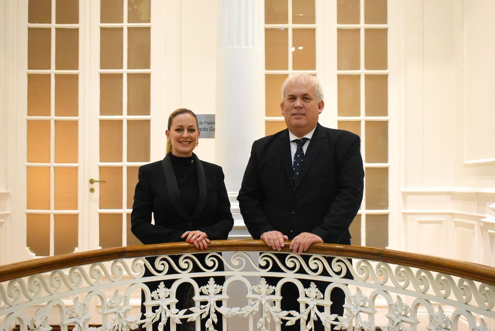 Dos autoridades del Consejo posan apoyadas en la baranda de hierro labrado de un balcón interior.