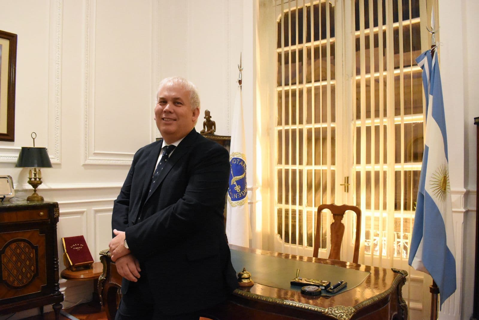 Una autoridad del Consejo en un despacho con escritorio de época, entre la bandera del notariado y la bandera argentina.